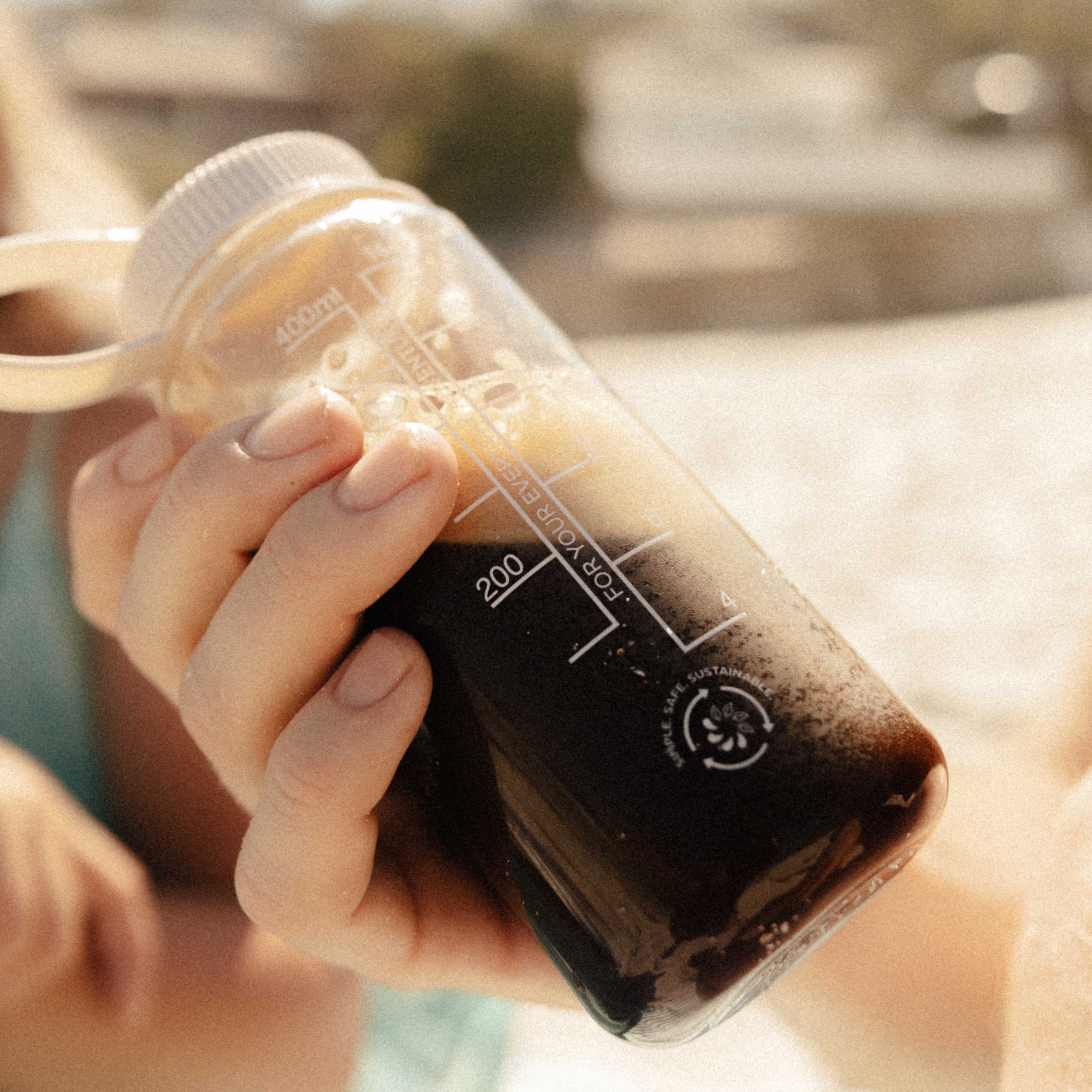 Hand holding a clear bottle with dark liquid against a blurred outdoor background