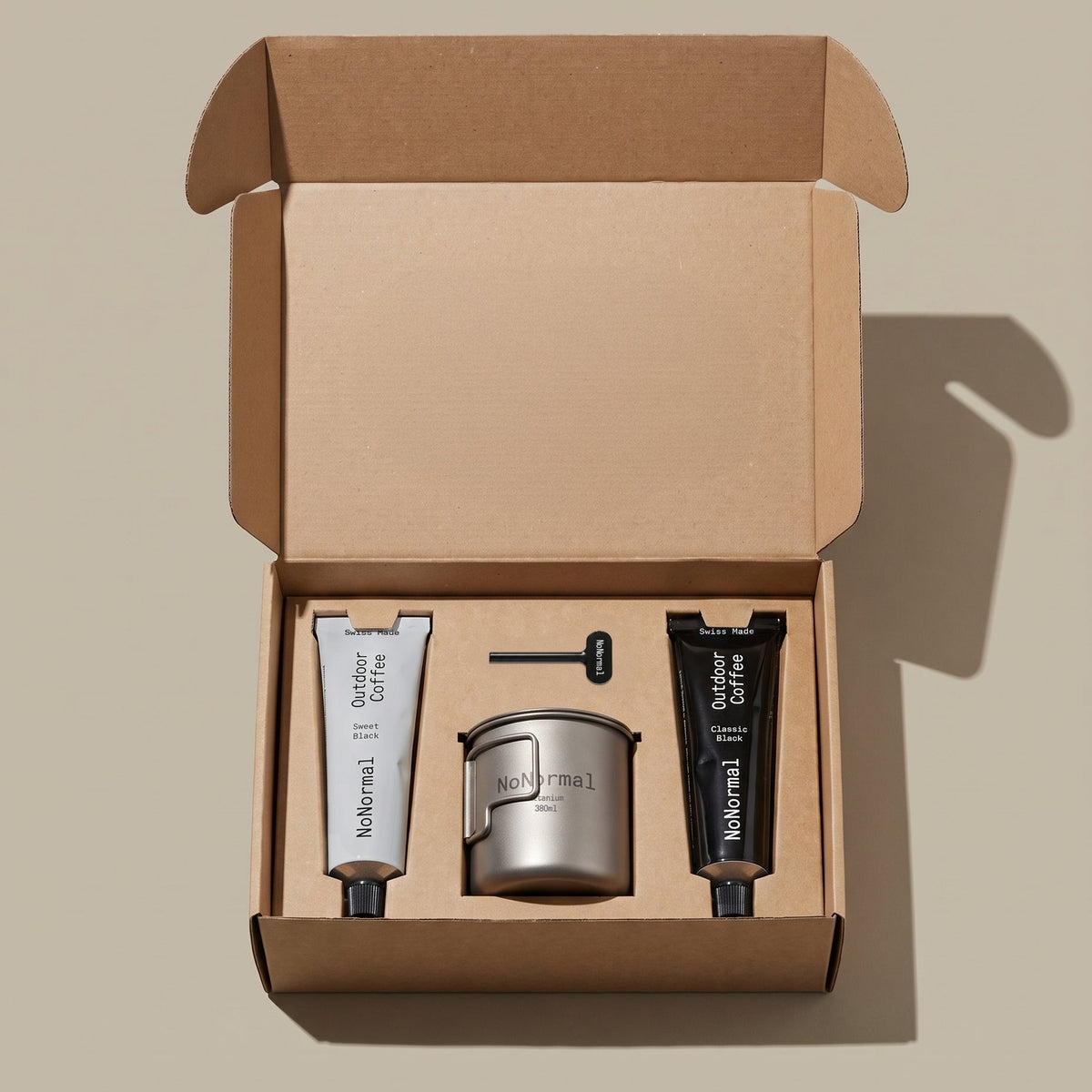 An open kraft cardboard gift box containing two nonormal coffee tubes a titanium cup and a tube squeezer
