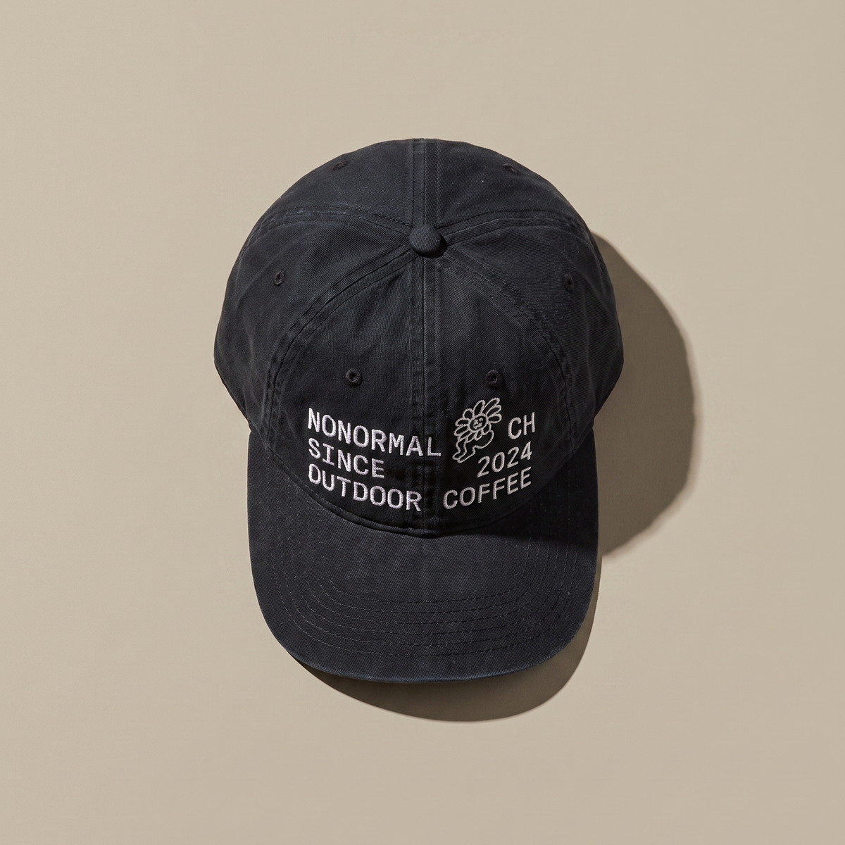 A washed black baseball cap seen from above on a beige background, with NONORMAL SINCE OUTDOOR COFFEE and CH 2024 embroidered in white across the front panel.