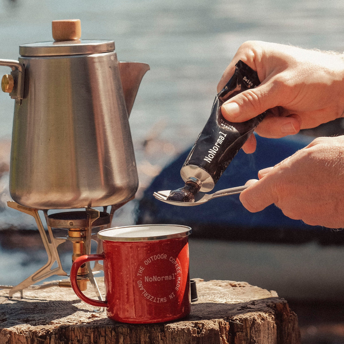 Squeezing NoNormal Classic Black onto a spoon beside a camping kettle at a mountain lake