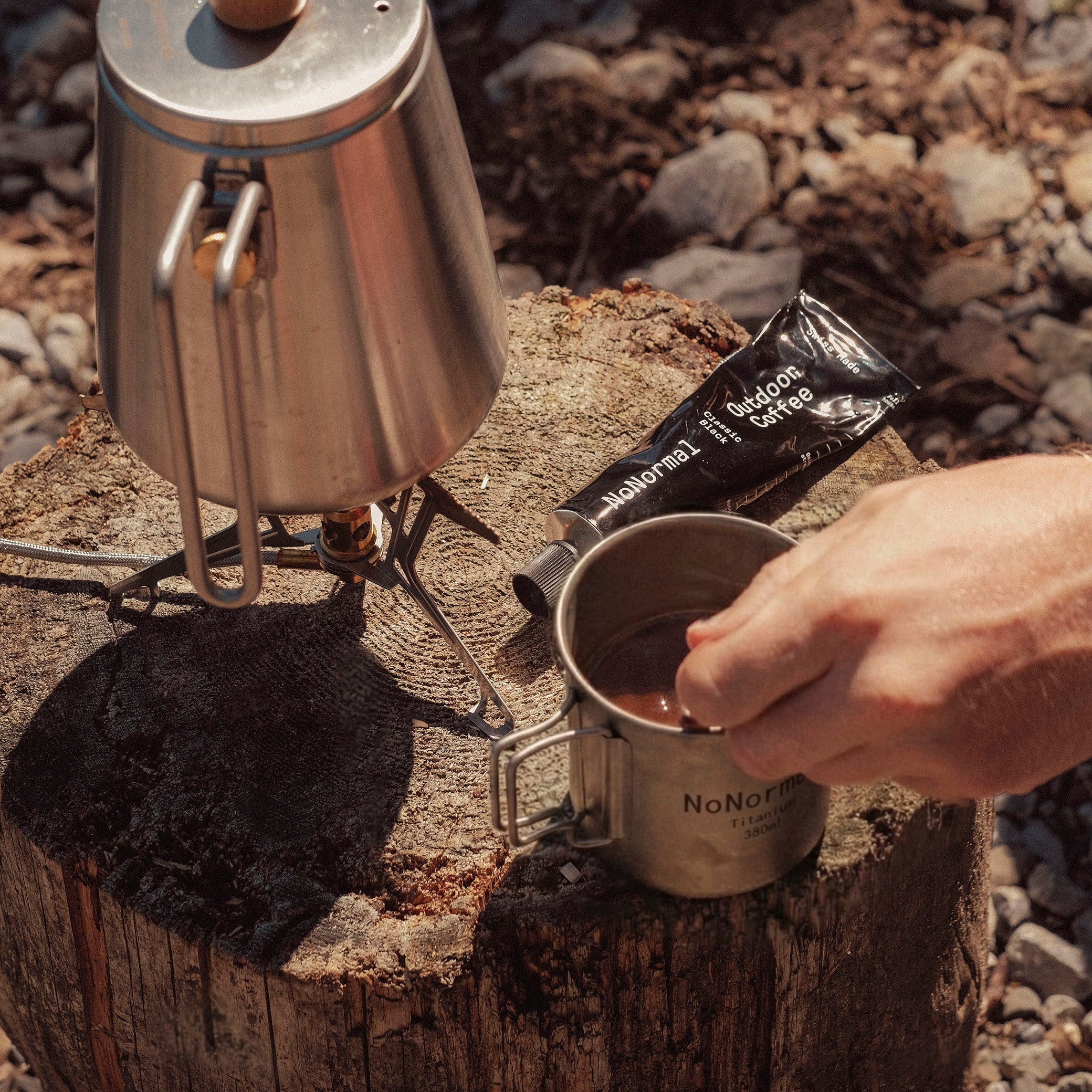 Camping coffee setup with a portable stove and mug on a log outdoors.