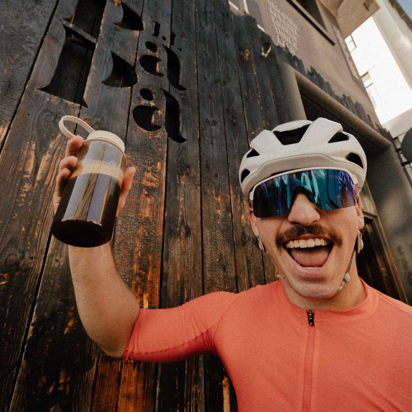 Person in cycling gear holding a water bottle, standing in front of a rustic wooden wall.