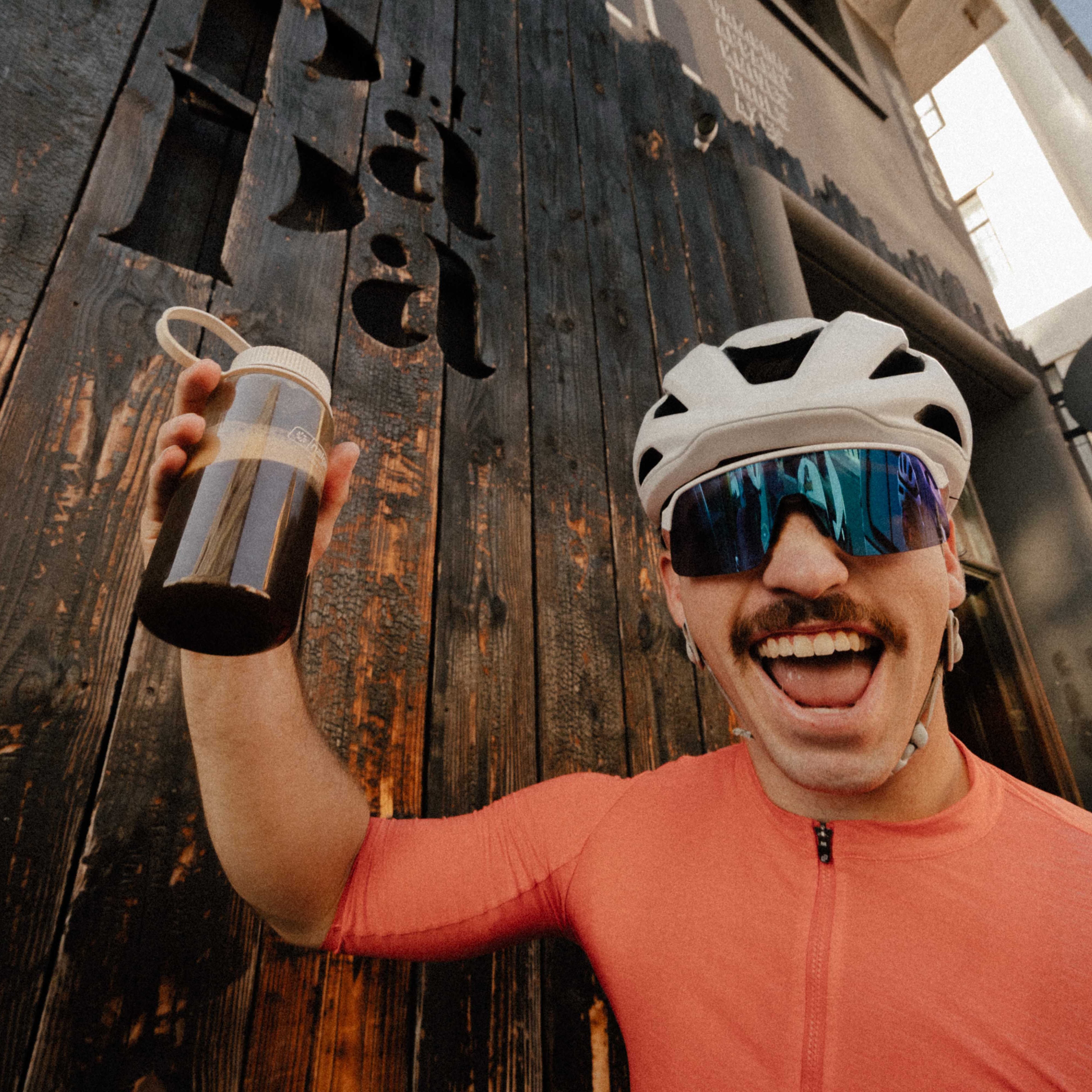 Person in cycling gear holding a water bottle, standing in front of a rustic wooden wall.