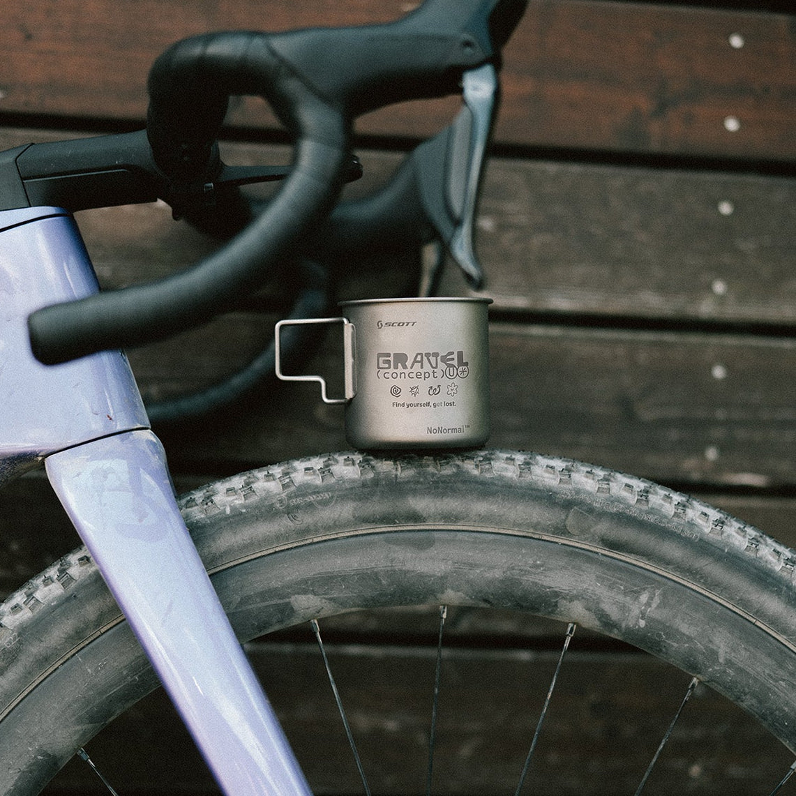 A close-up of a bicycle with a metallic silver cup mounted on the handlebar area, set against a dark wooden plank background.