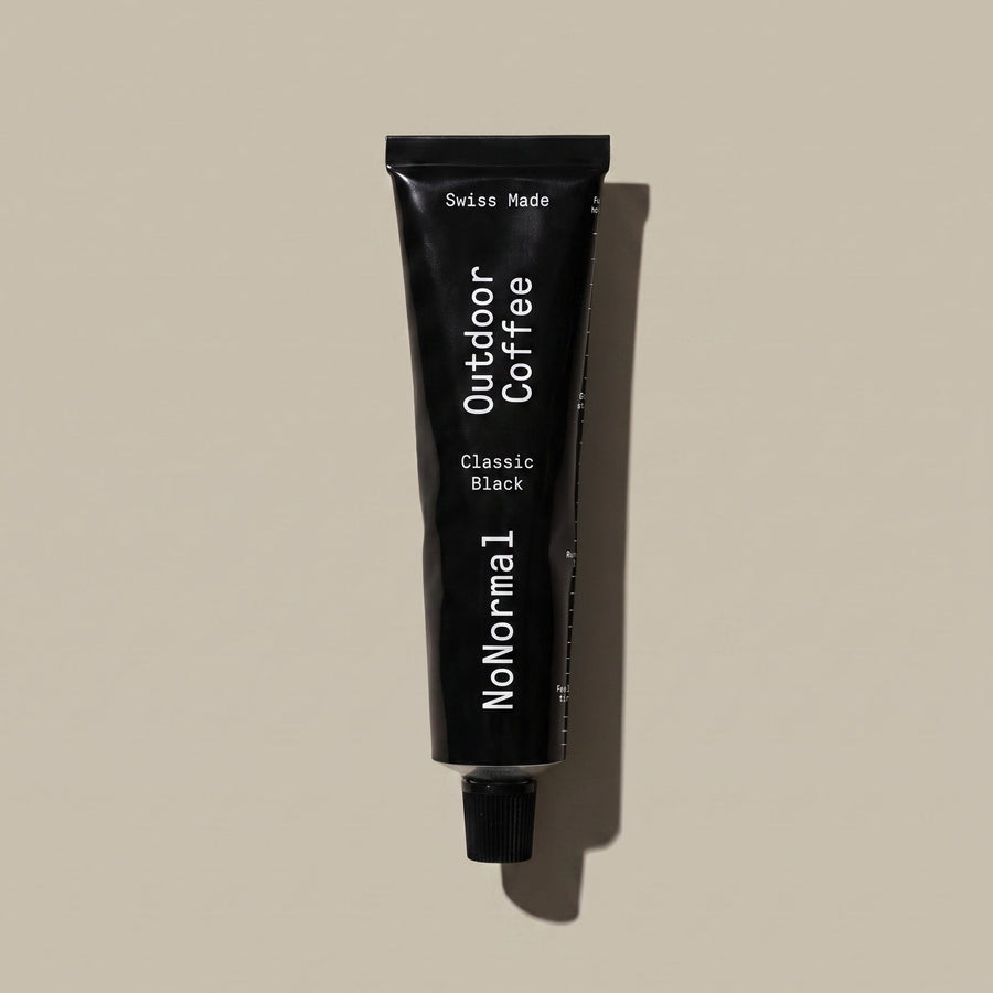 A black squeeze tube of NoNormal Outdoor Coffee Classic Black, standing vertically against a solid beige background with a soft shadow.