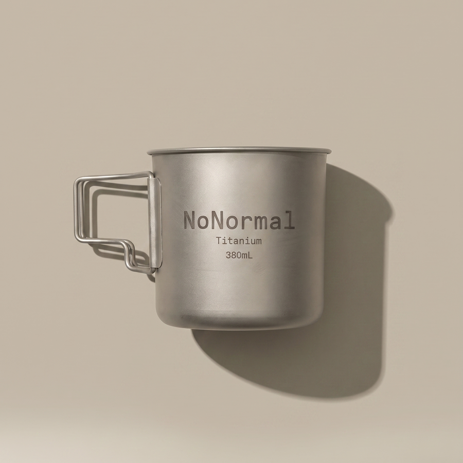 A titanium camping mug with a handle, featuring the text 'NoNormal Titanium 380mL' printed on its side, set against a solid beige background with a soft shadow.