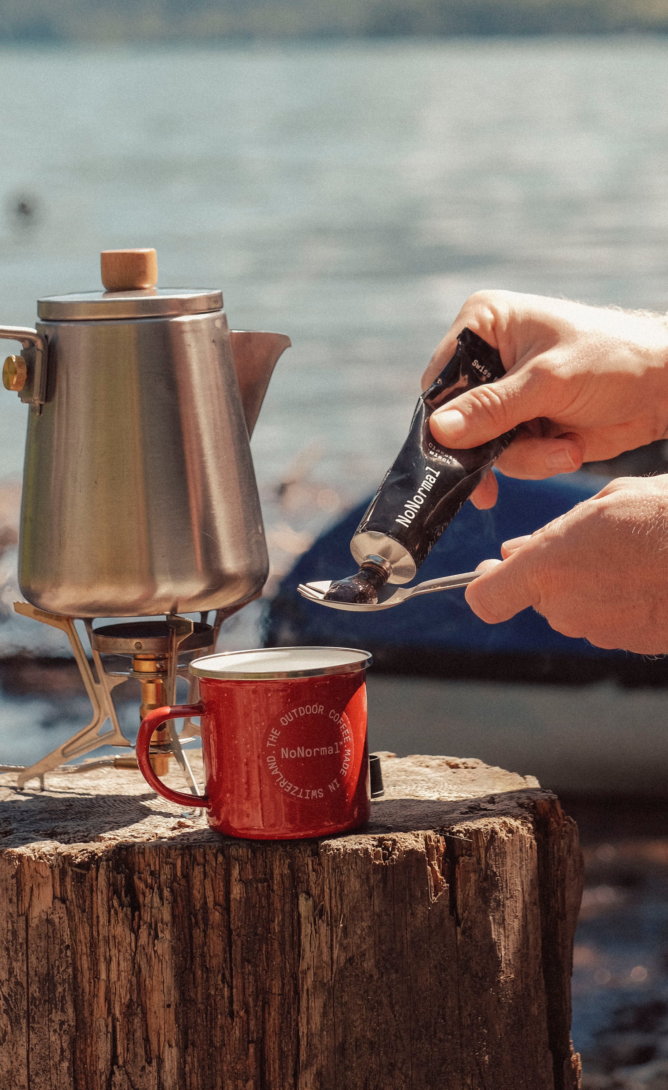 Squeezing NoNormal Classic Black onto a spoon beside a camping kettle at a mountain lake