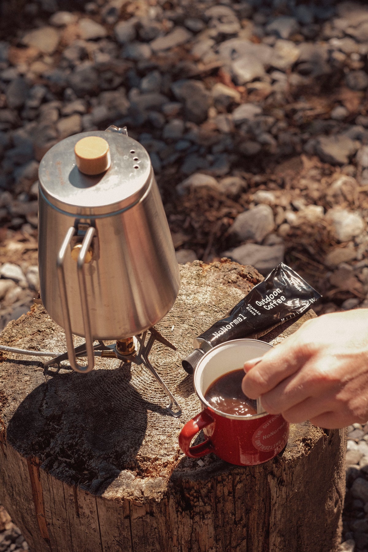 Stirring NoNormal Classic Black into a cup beside a camping kettle in the forest