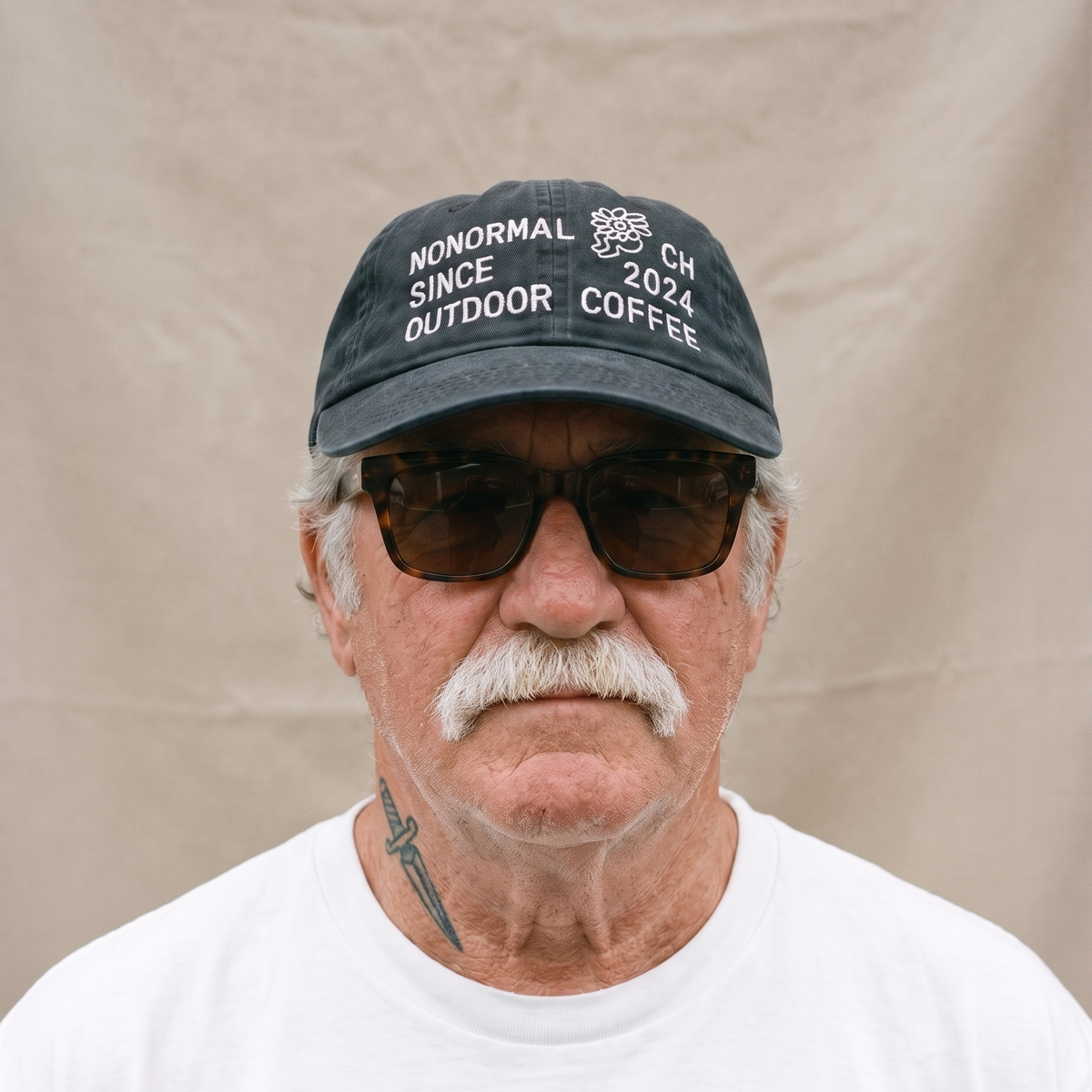 A close-up portrait of a man with a white mustache wearing a dark gray baseball cap that reads 'NONORMAL SINCE 2021 OUTDOOR COFFEE' and a white t-shirt, set against a neutral beige fabric backdrop.