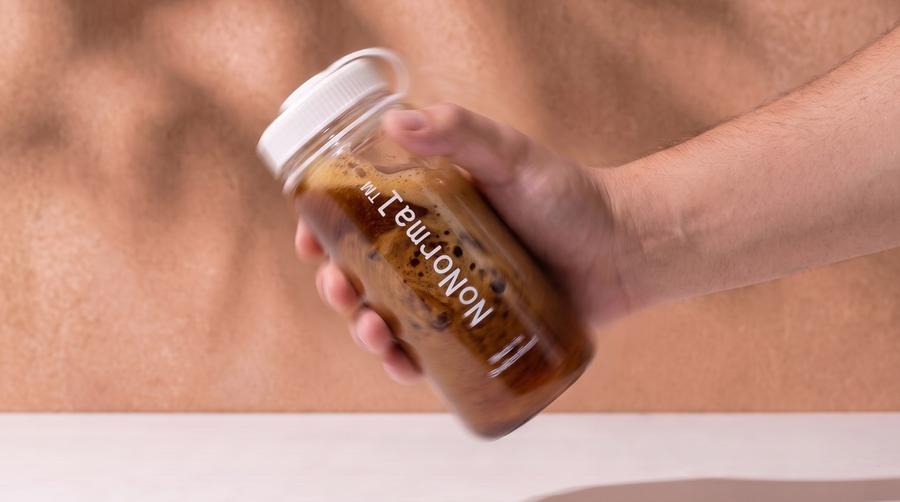 A hand holding a clear plastic jar with a white lid, filled with a brown beverage and featuring a white label that reads NoNormal'. The background is a warm, textured tan wall with soft leaf shadows, and a light surface below.