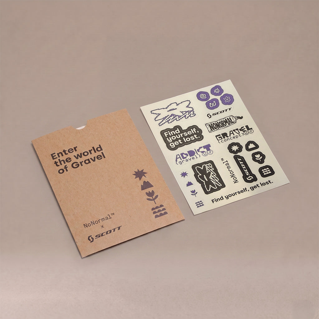 A kraft paper sleeve and a white sticker sheet featuring black and purple text stickers with motivational phrases, photographed on a neutral beige background. The sleeve displays 'Enter the world of Gravel' and 'NoNormal x SCOTT'.