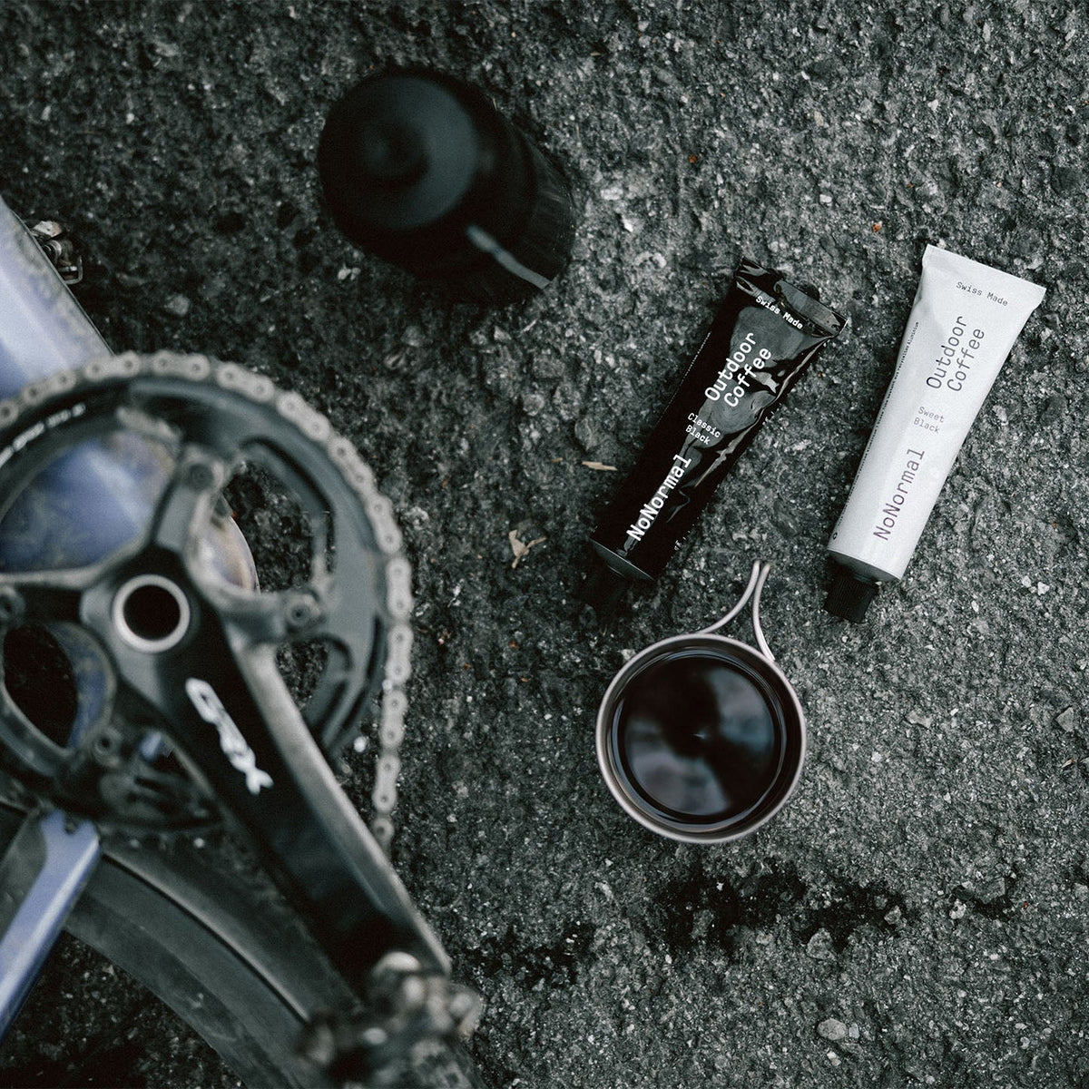 Bike chainstay and crankset laid on asphalt beside a tube of grease, a small cup of dark fluid, and a black bottle, photographed from above.