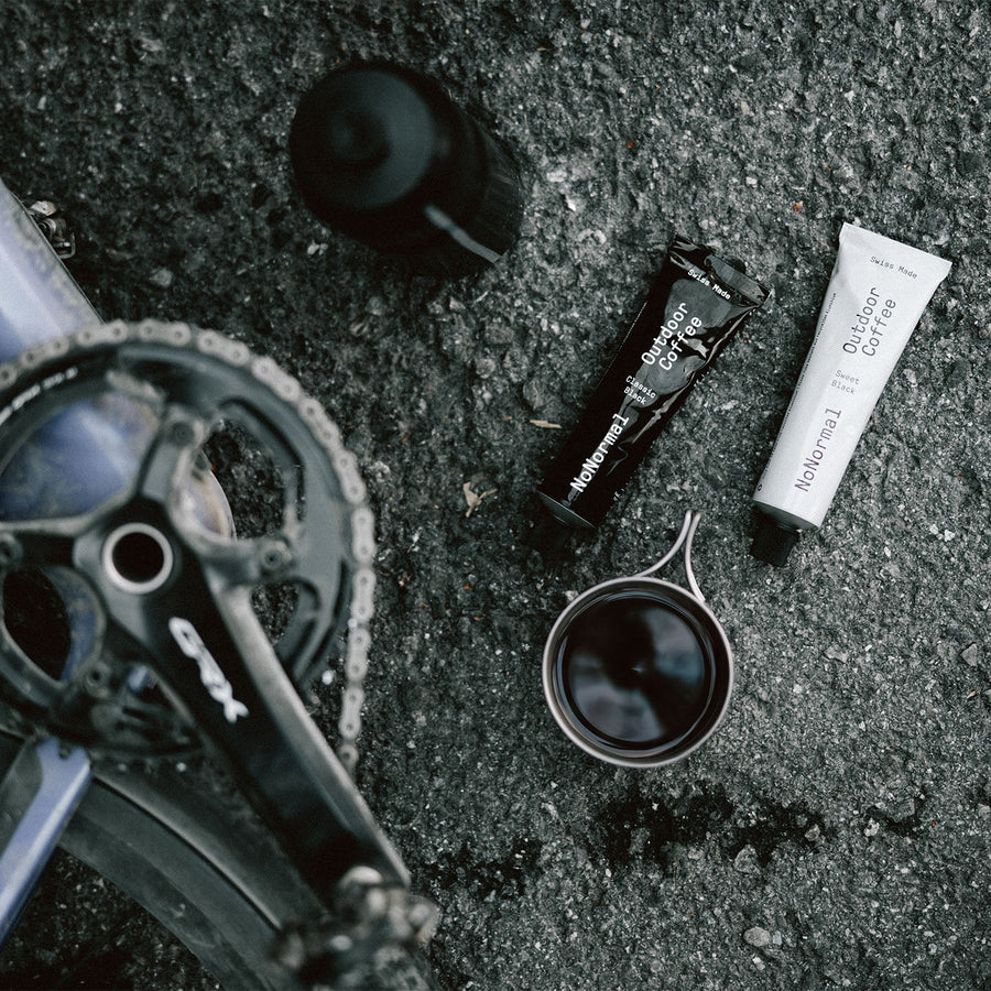 Bike chainstay and crankset laid on asphalt beside a tube of grease, a small cup of dark fluid, and a black bottle, photographed from above.