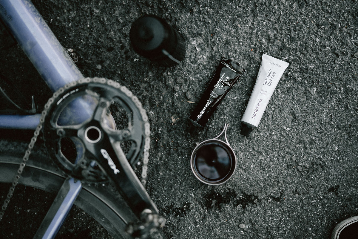 Bike chainstay and crankset laid on asphalt beside a tube of grease, a small cup of dark fluid, and a black bottle, photographed from above.
