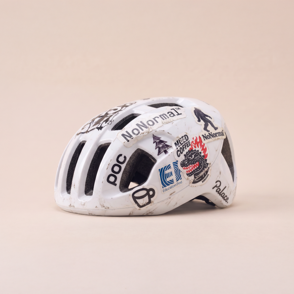 A white mountain bike helmet with various sponsor decals, including 'NoNormal', 'poc', and 'Palice', displayed on a solid beige background.
