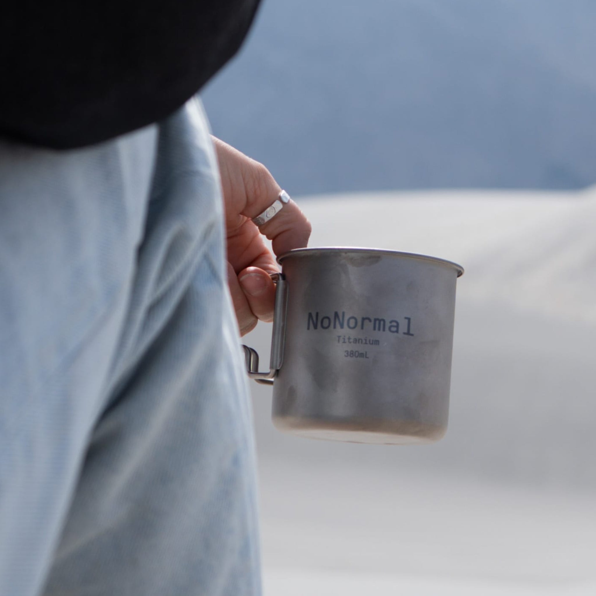 Person holding a metallic cup with 'NoNormal' branding against a blurred natural background